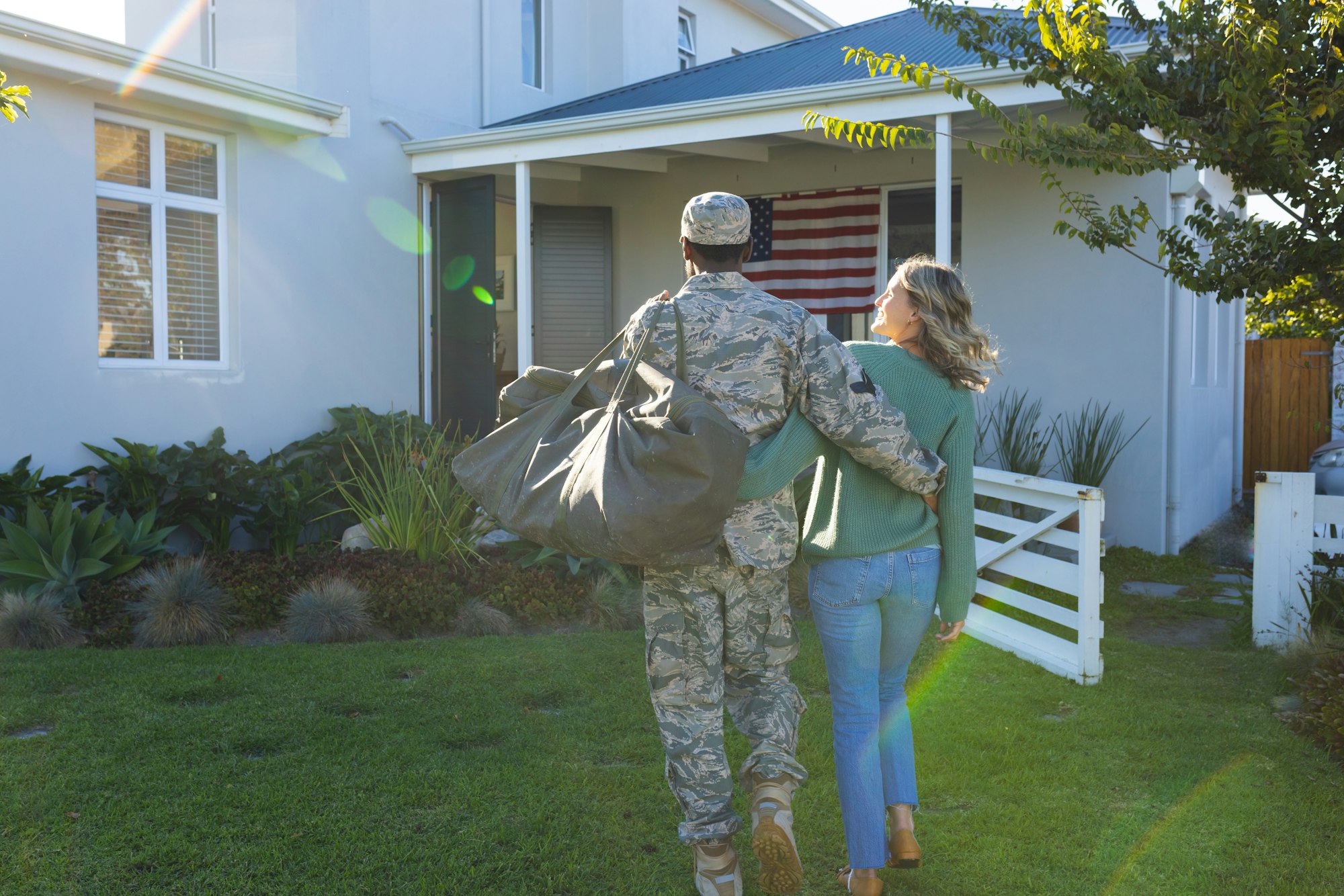 Rear view of solider with arm around multiracial woman carrying backpack and walking towards house